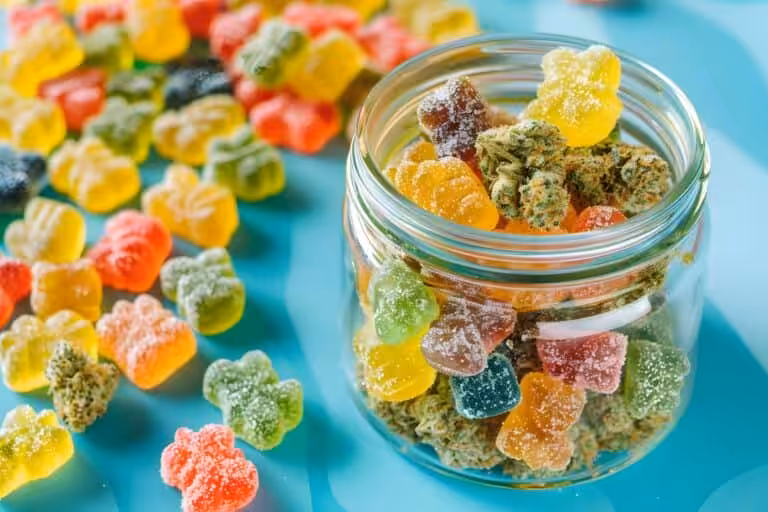 Bright, colorful cannabis gummies on a blue surface with a glass jar filled with assorted gummy candies and cannabis buds.